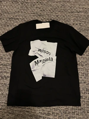 Maison Margiela T-Shirt - Svart t-shirt från Maison Margiela med korta ärmar och ett unikt vitt print framtill med skrynklat pappersmotiv och logga. Klassisk passform och rund halsringning. Perfekt för dig som gillar statement-plagg och designerstil.             🟥Kan gå ner i pris vid snabb affär 🟥