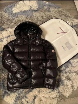 Svart Moncler pufferjacka med huva - Svar pufferjacka från Moncler med glansig finish och huva. Jackan har dragkedja framtill,användt den fåtal gånger hör av dig för minsta lila OBS passar storlek M också Kvitto och allt kommer med✅