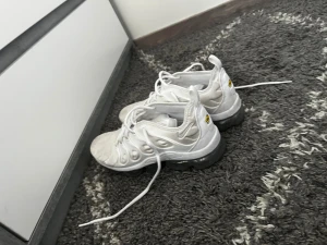 Nike Air Vapormax Plus vita sneakers - Nike Air Vapormax Plus sneakers i helvitt med genomskinlig sula och snygga detaljer på ovandelen. Skorna har snörning och en modern, sportig look med rund tå och platt sula. Perfekt för dig som gillar streetwear och vill ha något som sticker ut.