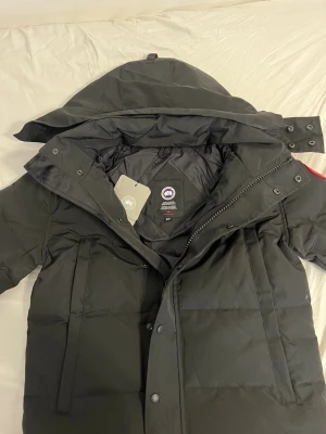 Svart dunjacka från Canada Goose - Svart dunjacka från Canada Goose med stor huva, klassisk logga på ärmen och praktiska fickor. Jackan har dragkedja och knappar framtill, quiltad design och är långärmad. Perfekt för kalla vinterdagar och har en clean, stilren look. PÄLS INGÅR SAMT DUSTBAG