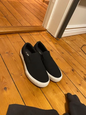 Svarta slip-on sneakers från 157 - Svarta slip-on sneakers från 157 med vit platt sula och klassisk rund tå. Skorna är tillverkade i textilmaterial och har elastiska paneler på sidorna för enkel på- och avtagning. Yttersulan är mönstrad i gummi för bra grepp. Perfekt för en chill och enkel stil. De har aldrig använts utomhus. 