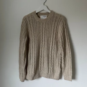 Beige kabelstickad tröja Selected Homme - Snygg beige kabelstickad tröja från Selected Homme i storlek M. Tröjan har rund hals, ribbade muddar och ett klassiskt kabelmönster som ger en chill och stilren vibe. Perfekt för lager-på-lager under hösten och vintern. 5H1