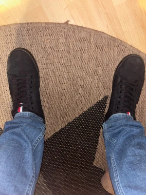 Svarta sneakers Tommy Hilfiger - Snygga svarta sneakers från Tommy Hilfiger med klassisk rund tå och platt sula. Skorna har snörning och detaljer i rött, vitt och blått på plösen och sidan. Materialet är canvas och de har en stilren, enkel design som passar till jeans och streetwear. Använda en gång 