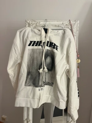 Vit the cure hoodie - Säljer en vit hoodie med The Cure-tryck fram och på ena ärmen. Tröjan har dragkedja, snörning vid huvan och ribbade muddar. Materialet är mjuk bomull och passformen är normal. Perfekt för dig som gillar bandmerch och streetstyle.