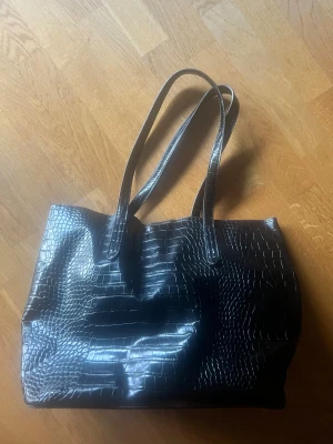 Svart shoppingväska i skinnimitation - Stilren svart shoppingväska med croco-mönstrad yta och långa axelremmar. Väskan har en rymlig form och är tillverkad i skinnimitation som ger en cool och modern vibe. Perfekt för dig som vill ha plats för allt du behöver under dagen.