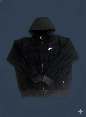 Svart Nike vindjacka med huva - Svart vindjacka från Nike i storlek M. Jackan har huva med snörning, dragkedja framtill och två sidofickor med dragkedja. Nike-logga i vitt på bröstet. Tillverkad i lätt polyester, perfekt för blåsiga dagar. Stilren och sportig look.