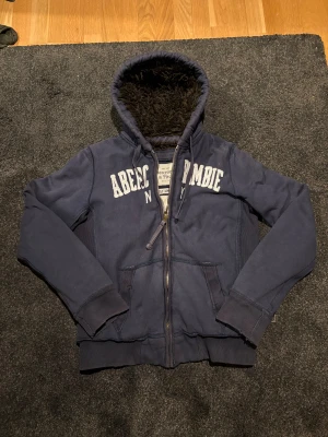 Abercrombie & Fitch Pälscardigan - Mysig mörkblå hoodiejacka från Abercrombie & Fitch med stor vit logga på bröstet. Jackan har dragkedja, två fickor fram och en fluffig brun fuskpäls i huvan. Perfekt för kalla dagar när du vill vara både varm och snygg.