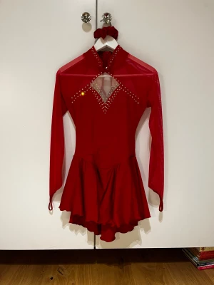 Röd glittrig konståkningskläning  - Snygg röd konståkningskläning med långärmade meshärmar och glittriga stenar framtill. Klänningen har en kort, volangig kjol och en cool öppen rygg med korsade band. Perfekt för dig som vill sticka utt! Scrunchie i matchande färg ingår.