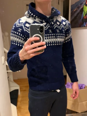 Mörkblå stickad hoodie med mönster - Säljer en sjukt snygg mörkblå stickad hoodie med vitt och grått nordiskt mönster över axlar och bröst. Tröjan har huva med snörning och långa ärmar. Perfekt för kalla dagar och har en riktigt skön och mysig känsla. Passar dig som gillar klassisk vinterstil. Han på bilden är 178 cm lång och upplever att den är för liten😇