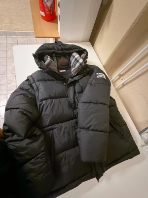 Svart pufferjacka från Burberry - Säljer en svart pufferjacka från Burberry med avtagbara ärmar så den kan användas som väst. Jackan har huva med rutigt foder i grått, vitt och rött samt Burberry London England-patch på ärmen. Stora fickor med dragkedja och knappar framtill. Perfekt för kalla dagar. PRIS KAN DISKUTERAS ÄNDA SERIÖSA KÖPARE