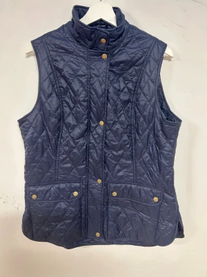Mörkblå quiltad väst från Barbour - Quiltad mörkblå väst från Barbour med hög krage, dragkedja och tryckknappar. Västen har två stora fickor framtill och klassiskt rutmönster i tyget. Perfekt för lager-på-lager och snygg till både jeans och boots.