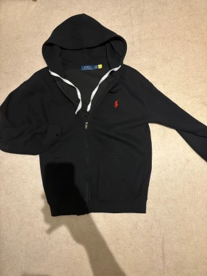 Svart hoodie från Polo Ralph Lauren - Svart hoodie från Polo Ralph Lauren med röd broderad logga på bröstet. Tröjan har huva med grå snörning, dragkedja framtill och två fickor. Tillverkad i mjuk bomull, perfekt för chill dagar. Klassisk och stilren design.