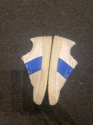 Valentino Garavani vita sneakers med blå detalj - Säljer ett par vita Valentino Garavani sneakers med en bred blå rem över sidan. Skorna har rund tå, platt sula och klassisk snörning. Tillverkade i skinn för en lyxig känsla och stilren look. Perfekta för dig som vill sticka ut med designerstil och perfekta för den uppkommande vintern. Jag måste bara säga att skorna har ett hål i sulan på skorna. Jag rengör såklart skorna inannan dem skickas.
