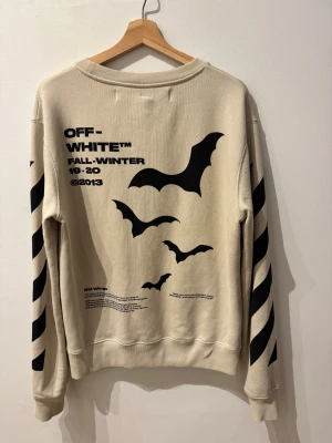 Beige Off-White sweatshirt med batprint - Snygg beige sweatshirt från Off-White med svarta bat-prints på framsidan och coola diagonala ränder på ärmarna. Crewneck och ribbade muddar. Perfekt för dig som gillar streetwear och vill sticka ut med en unik design från Virgil Abloh.