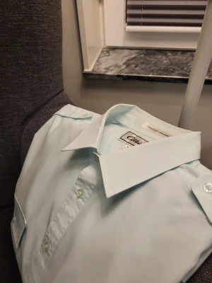 Ljusblå kortärmad skjorta Atkins L - Kortärmad ljusblå skjorta från Atkins med klassisk krage, två bröstfickor med lock och knappar. Skjortan är tillverkad i en mix av polyester och bomull, perfekt för en clean och stilren look. Snygga axelpartier med knappdetaljer.