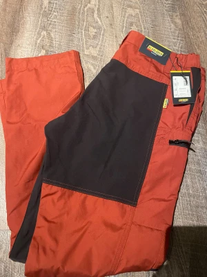 Röd-svarta Blåkläder byxor C46 - Snygga röd-svarta byxor från Blåkläder, modell 7159. Byxorna har en cool färgblockad design med svarta förstärkningar på framsidan och praktisk dragkedja på benet. Perfekta för dig som gillar funktion och stil. Materialet är slitstarkt och passar för aktiva dagar.