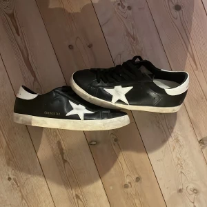 Svarta Golden Goose Superstar sneakers !!!BYTE FUNKAR!!! - Golden Goose Superstar sneakers i svart skinn med vit stjärndetalj på sidan och vit häl. Klassisk låg modell med snörning och platt sula. Märkets logga syns på sidan och plösen. Perfekta för en cool och avslappnad stil. Knappt andvöndna 
