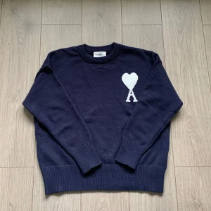 Ami tröja - Snygg mörkblå stickad tröja från Ami med vit hjärtlogga på bröstet. Klassisk rund halsringning, ribbade muddar och lång ärm. Perfekt för dig som gillar stilrena plagg med en twist. Passar till jeans eller dressade byxor. 