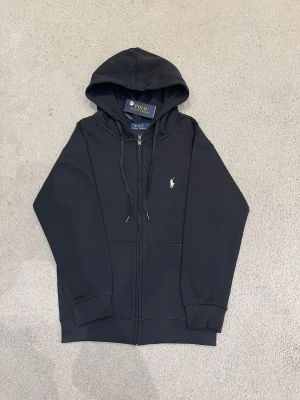 Ralph Lauren Kofta  - Svart hoodie från Polo Ralph Lauren med dragkedja och snörning i huvan. Klassisk design med vit broderad logga på bröstet. Tillverkad i mjuk bomull och har långa ärmar samt ribbade muddar vid ärmslut och nederkant.