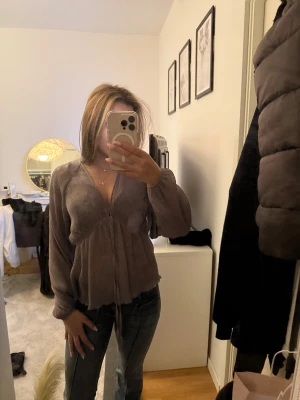 Brun omlott blus med knyt - Säljer en brun blus med omlottkänsla och knyt framtill. Blusen har långa, lite puffiga ärmar och är tillverkad i ett lätt, ribbat material. Den har v-ringning och en flowy passform som ger en avslappnad men ändå trendig look.