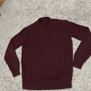 Vinröd stickad tröja Ralph Lauren - Säljer en vinröd stickad tröja från Polo Ralph Lauren i storlek XS. Tröjan har rund halsringning, ribbade muddar och det klassiska gröna broderade logot på bröstet. Perfekt för dig som gillar stilrena och tidlösa plagg.