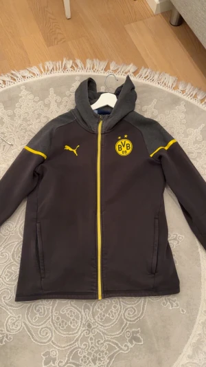Borussia Dortmund hoodiejacka Puma - Mörkgrå hoodiejacka från Puma med Borussia Dortmund-logga och gula detaljer. Jackan har huva, hel dragkedja i gult och två sidofickor. Materialet är mjukt och känns som bomull eller polyester. Perfekt för dig som gillar fotboll och vill ha en sportig look.