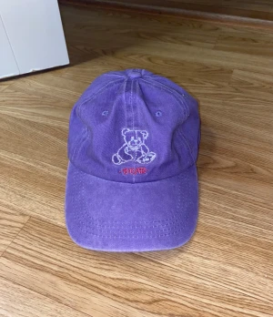 Lila keps med nallebrodyr - Säljer en lila keps med broderad nalle och texten 'BEAR' framtill. Kepsen har böjd skärm, justerbar rem baktill och är tillverkad i bomull.  Använder aldrig bara legat i garderoben 
