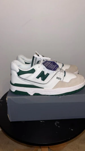 New Balance 550 vit/grön sneakers - Säljer ett par New Balance 550 sneakers i vitt och mörkgrönt med snygga detaljer. Skorna har en platt sula, rund tå och snörning. Ovandelen är i skinn och mocka med meshpaneler för extra komfort. Ikonisk N-logga på sidan och 550-tryck. Perfekt för dig som gillar retrostil.