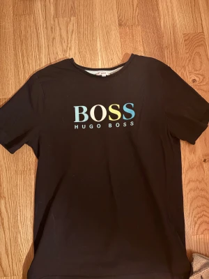 Svart BOSS t-shirt med färgglad logga - Svart t-shirt från Hugo Boss i storlek XS med rund hals och korta ärmar. Framsidan har en stor BOSS-logga i blått, gult och ljusblått. Materialet är mjuk bomull och passformen är normal. Perfekt för dig som gillar stilrena märkesplagg med tydlig branding.