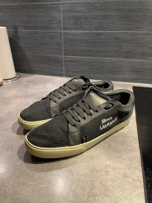 Svarta Saint Laurent sneakers - Svarta sneakers från Saint Laurent med broderad logga på sidan och guldfärgad text på plösen. Skorna har snörning, rund tå och en beige platt sula. Ovandelen är i canvas med detaljer i skinn, vilket ger en sjysst kontrast. PRIS EJ HUGGET I STEN VID SNABB AFFÄR 