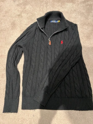 Svart stickad polotröja Polo Ralph Lauren - Svart kabelstickad polotröja från Polo Ralph Lauren med half zip och röd broderad logga på bröstet. Tröjan har ribbade muddar och hög krage, perfekt för kyliga dagar. Klassisk och stilren design som funkar till jeans eller chinos.