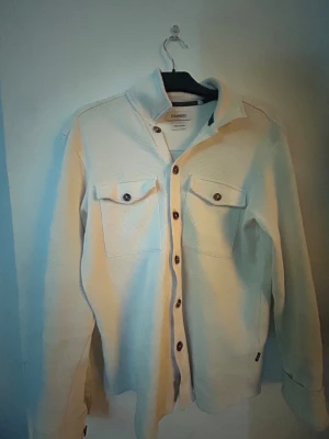 Vit overshirt från Strömberg - Vit overshirt från Strömberg i bomull.  klassisk krage och bruna knappar längs hela framsidan. Perfekt lager-på-lager-plagg med en fräsch slutlook priset på tröjan är inte bestämt då ja inte vet vad en sån kostar, så hör av er :)