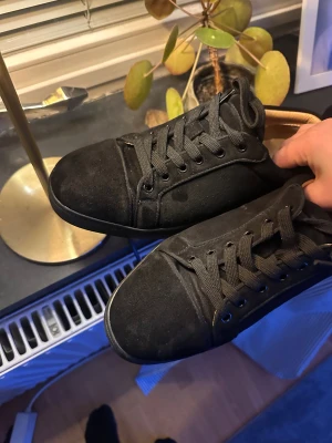 Svarta mocka sneakers från Louboutin - Säljer ett par svarta sneakers från Christian Louboutin i mjuk mocka med klassisk röd sula och rund tå. Skorna har snörning och en stilren, platt sula. Insidan är beige och har Louboutin-loggan. Perfekt för dig som gillar lyxiga detaljer och clean look.