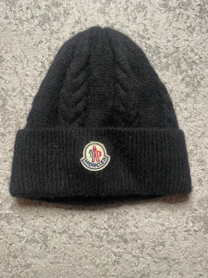 Svart Moncler kabelstickad mössa - Svart kabelstickad mössa från Moncler med bred uppvikt kant och klassisk Moncler-logga framtill. Mössan är tillverkad i mjuk ull och har ett stilrent, chunky flätmönster som ger extra vibe till din vinterstil.