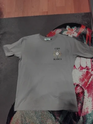 Grå t-shirt från Casablanca - Snygg grå t-shirt från Casablanca med tryck på bröstet där det står 'Casa Blanca' och en dekorativ emblem. Klassisk passform, rund hals och tillverkad i mjuk bomull. Perfekt för en clean och stilren look.