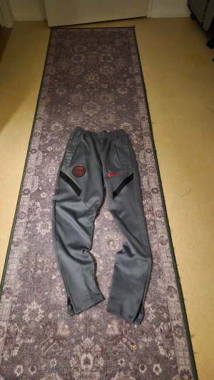 PSG x Nike grå träningsbyxor - Grå träningsbyxor från Nike med PSG-logga och röd swoosh på framsidan. Byxorna har elastisk midja, dragkedjor vid bensluten och svarta detaljer längs benen. Perfekta för fotboll eller chill, med sportig look och bekväm passform.