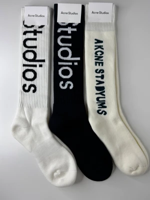 Acne Studios vita och svarta strumpor - Tre par ribbade strumpor från Acne Studios i svart och vitt med stor logotyp längs skaftet. Tillverkade i mjuk bomullsblandning med 90% bomull, 8% polyamid och 2% elastan. Perfekta för att ge din outfit en clean och trendig touch.