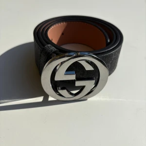 Svart Gucci bälte med GG-spänne - Säljer ett svart Gucci bälte med klassiskt GG-mönster och en stor, rund silverfärgad GG-logga som spänne. Bältet är tillverkat i skinn med en snygg mörk finish och detaljerad struktur. Insidan är beige och har präglad Gucci-text. Perfekt för dig som vill ha en lyxig accessoar.