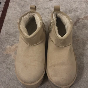 Beige boots med plattform och foder - Säljer ett par beige boots med platt sula och plattform. Skorna har rund tå och är fodrade med mjukt syntetpäls för extra värme. Utsidan är i mocka och de har dekorativa band på sidan. Perfekta för kalla dagar och har en enkel, stilren look.