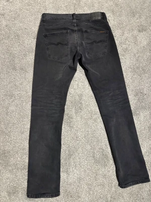 Nudie Jeans grim Tim - Säljer ett par svarta jeans från Nudie Jeans i storlek W31 L32. Jeansen har klassisk femficksdesign, svarta sömmar och Nudie Jeans logga på bakfickan. Materialet är mjukt denim. Model grim Tim.