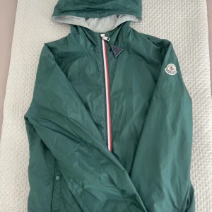 Grön vindjacka från Moncler  - Snygg grön vindjacka från Moncler med huva och dragkedja framtill. Jackan har Moncler-logga på ärmen och insida i grått tyg. Perfekt för dig som vill ha en stilren och sportig look. Klassisk passform och diskreta detaljer. Självklart äkta! Kvitto medföljer! NFC scannen funkar också. Går ner i pris vid snabb affär! Hör av dig vid minsta fundering. ☺️