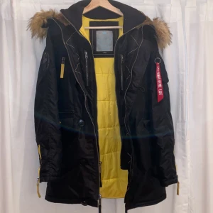 Svart parkajacka med päls från Alpha Industries - Säljer en svart parkajacka från Alpha Industries med gul insida och avtagbar päls på huvan. Jackan har dragkedja, knappar och flera fickor samt en ikonisk röd tag på ärmen. Perfekt för kyliga dagar.
