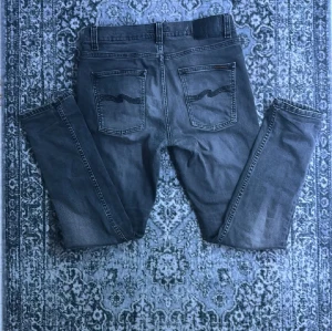 Grå Nudie Jeans Lean Dean 31/30 - Snygga grå jeans från Nudie Jeans, modellen Lean Dean. De har klassisk femficksdesign, svarta broderade vågor på bakfickorna och en smal passform. Materialet är 99% ekologisk bomull och 1% elastan för extra stretch. Lagning i skrevet som ej syns när man har på sig de (bild 4). Perfekt för dig som gillar stilrena och bekväma jeans.