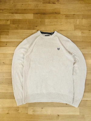 Vit stickad tröja från Chaps - Säljer en stilren vit stickad tröja från Chaps i 100% bomull. Storlek M.Tröjan har rund halsringning med svart detalj på insidan och en broderad logga på bröstet. Perfekt för dig som gillar klassisk och enkel stil.