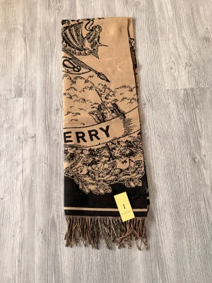 Burberry halsduk i kashmir med rutor och riddarmotiv - Exklusiv halsduk från Burberry i kashmir, med klassiskt rutig mönster på ena sidan och ett detaljerat riddarmotiv med texten 'BURBERRY' på den andra. Färgerna går i beige, brun och svart med inslag av rött. Långa fransar i ändarna ger en cool touch.