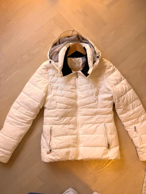 Vit pufferjacka från Norrsken - Snygg vit pufferjacka från Norrsken med huva och dragkedja framtill. Jackan har quiltad design, två fickor med dragkedja och justerbar huva. Perfekt för kalla dagar och har en clean look som funkar till många stilar. Sminkfläck på krage, går bort i tvätt