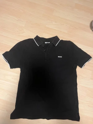 Svart kortärmad piké från Hugo boss - Svart pikétröja från Hugo boss i storlek xs/s med vit kant på krage och ärmslut. Klassisk design med tre knappar framtill och diskret BOSS-logga på bröstet. Perfekt för dig som gillar stilrena och enkla plagg med snygga detaljer.