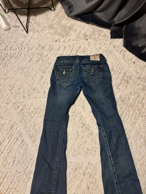 True Religion blå jeans med broderade fickor - Säljer ett par snygga blå jeans från True Religion med ikoniska broderade bakfickor och silverfärgade. Rätt så bra skick men har ett litet hål mellan benen men har gjort ett stygn