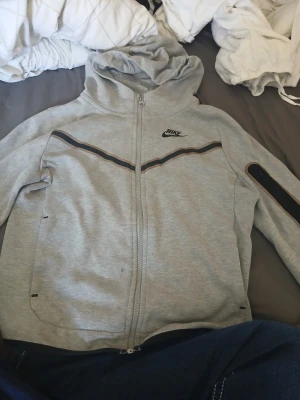 Grå hoodie från Nike med dragkedja - Snygg grå hoodie från Nike i storlek M, med hel dragkedja framtill och huva. Tröjan har svarta och bruna detaljer längs bröstet och på ena ärmen, samt Nike-logga på vänster sida. Perfekt för chill eller träning.