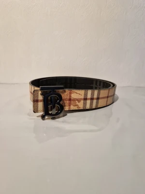 Rutig Burberry bälte med B-spänne - Snyggt bälte från Burberry med klassiskt rutigt mönster i beige, brunt och rött. Bältet har ett stort svart B-spänne framtill som ger en cool statement-look. Perfekt accessoar för att lyfta din outfit.